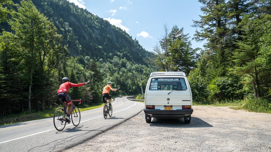 Two cyclists pass a white wilderness van on the road.