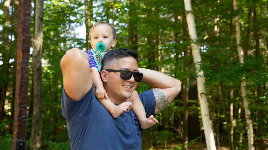 A man holds his baby on his shoulders while walking a nature path. 