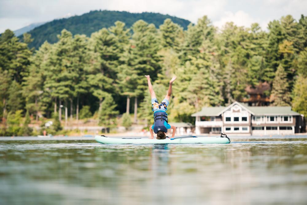 A teenager in swim clothing and a life jacket does a handstand on a stand-up paddleboard on a lake.