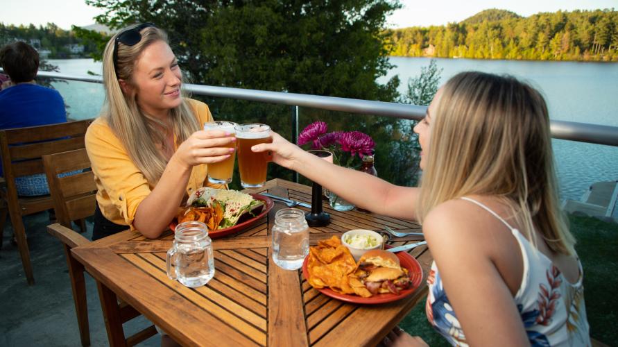 Two women toast glasses of beer at restaurant table overlooking a scenic lake.