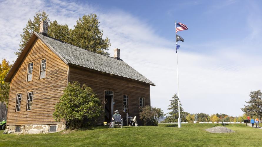 A nineteenth century farmhouse amid a sunny blue sky, with flags flying on a flagpole. 