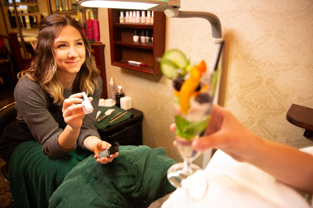A woman offers nail polish selections to a spa client enjoying a fruit dessert.