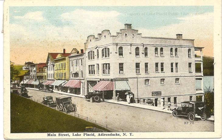 Vintage postcard of a row of buildings in Lake Placid, with early autos on the street. Image courtesy Lake Placid Public Library.