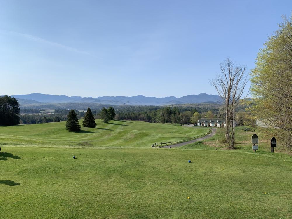 Tee and fairway at Lake Placid Club golf course, with Olympic ski jumps and Adirondack peaks in the distance.