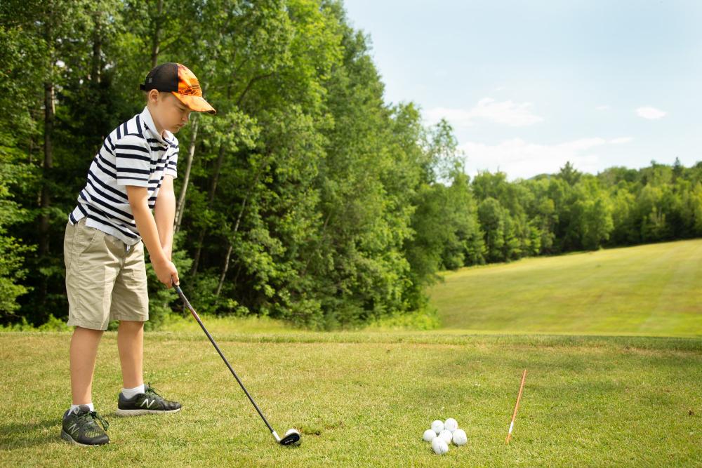 A young boy prepares to tee off on a golf green, with bright green trees along the fairway.