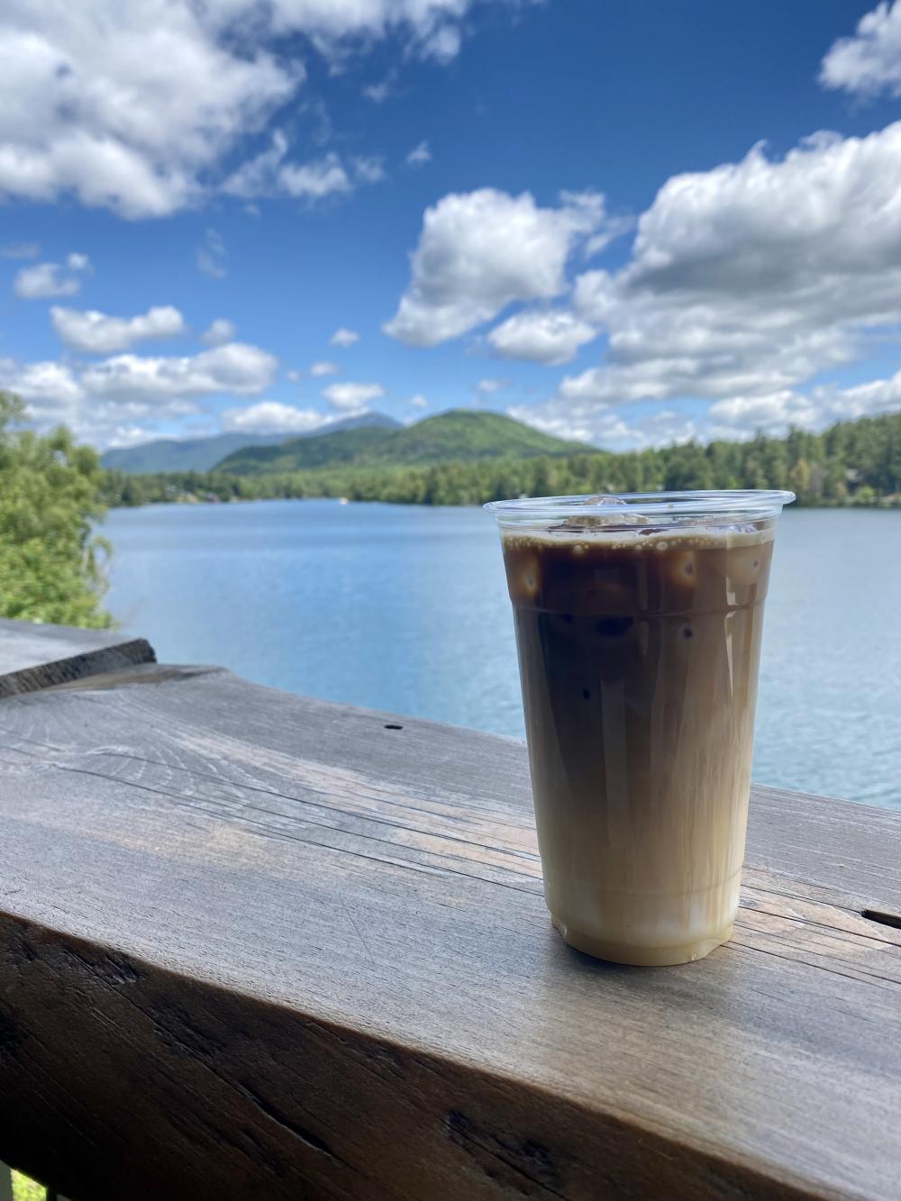 A cold coffee sits on a railing overlooking a sunny lake and mountains.