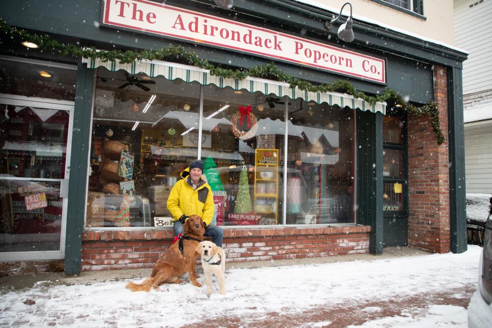 A man with two dogs sitting outside Adirondack Popcorn Co. decorated for the holidays.
