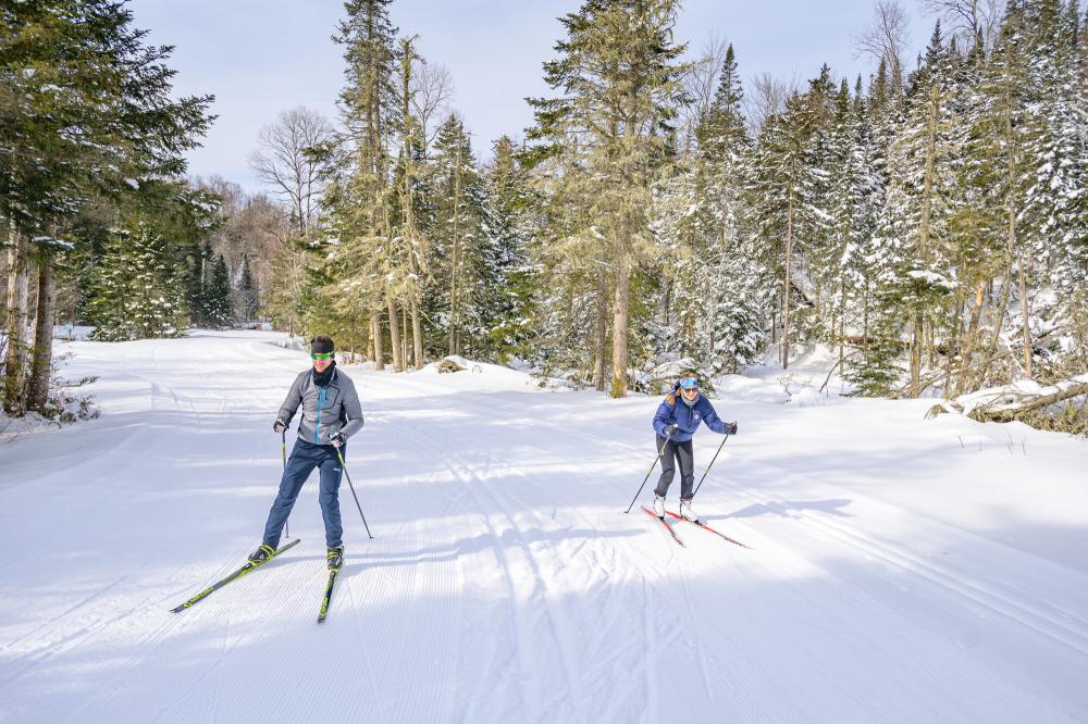 A man and woman ski on a groomed trail at a ski center with pine trees in the background.