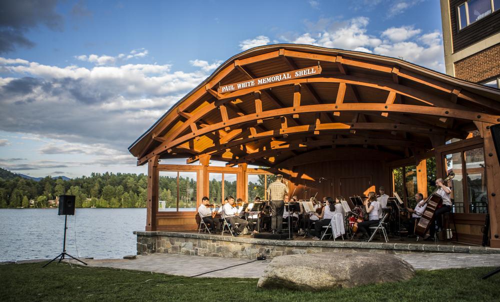 A sinfonietta playing instruments on a bandshell stage with a lake in the background.