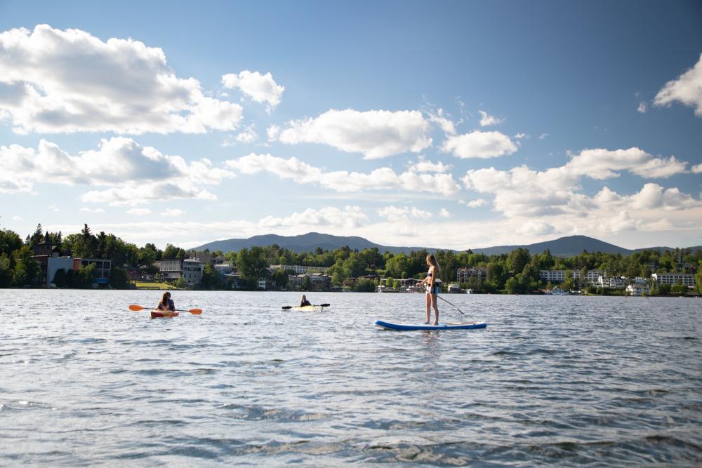 Kayakers and a woman on a SUP relax on a lake with town buildings in the background.