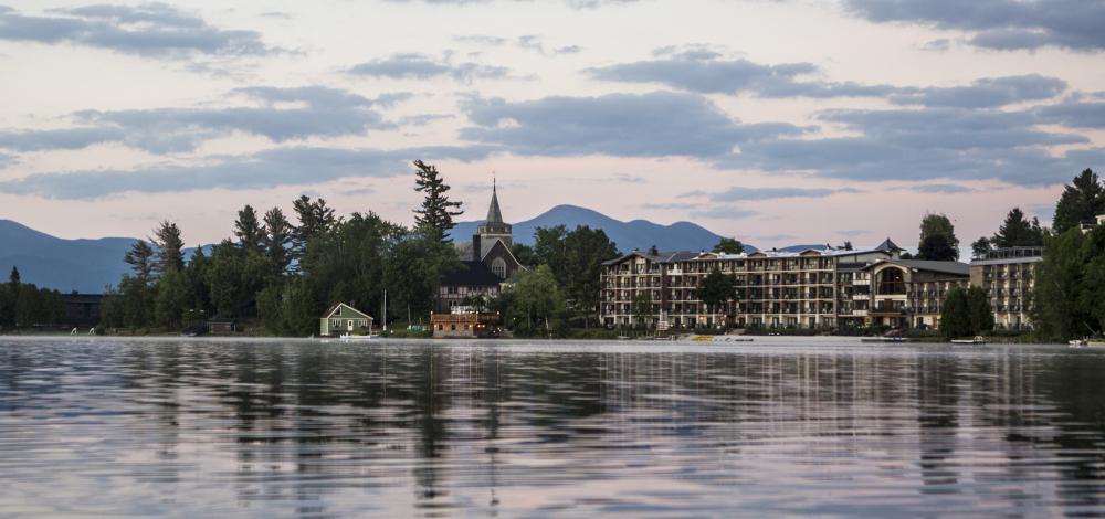A wide hotel, church, and other buildings amid trees and a backdrop of mountains with a lake in the foreground.