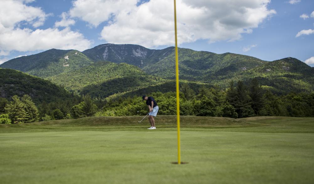 A man hits a golf ball into a hole with mountains in the background.