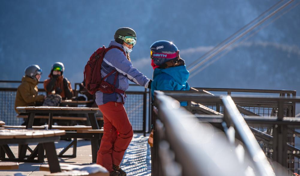 Two people talk outdoors on a patio dressed in ski gear, with helmets and jackets and snowpants.