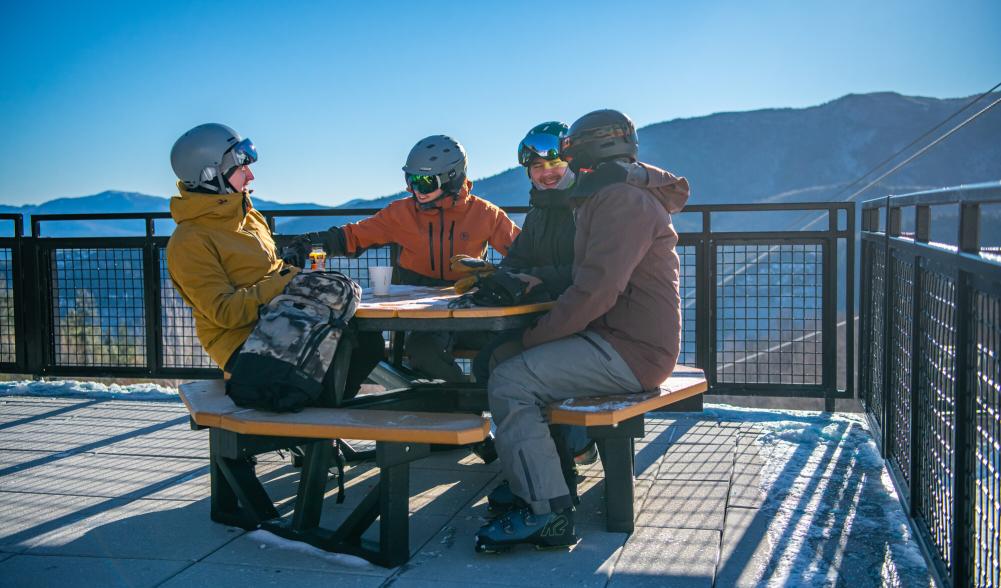 Four friends smile and laugh at an outdoor picnic table on a patio while dressed in ski clothes.
