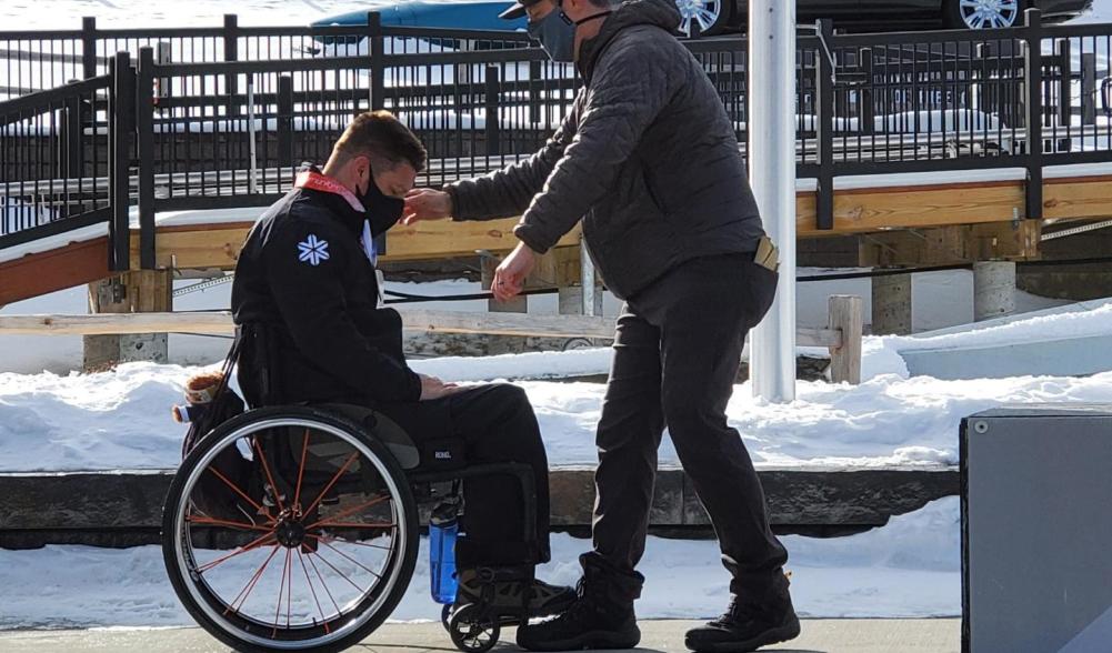 A man stands beside a man in a wheelchair to help him along a sidewalk. Image courtesy of David Christopher.