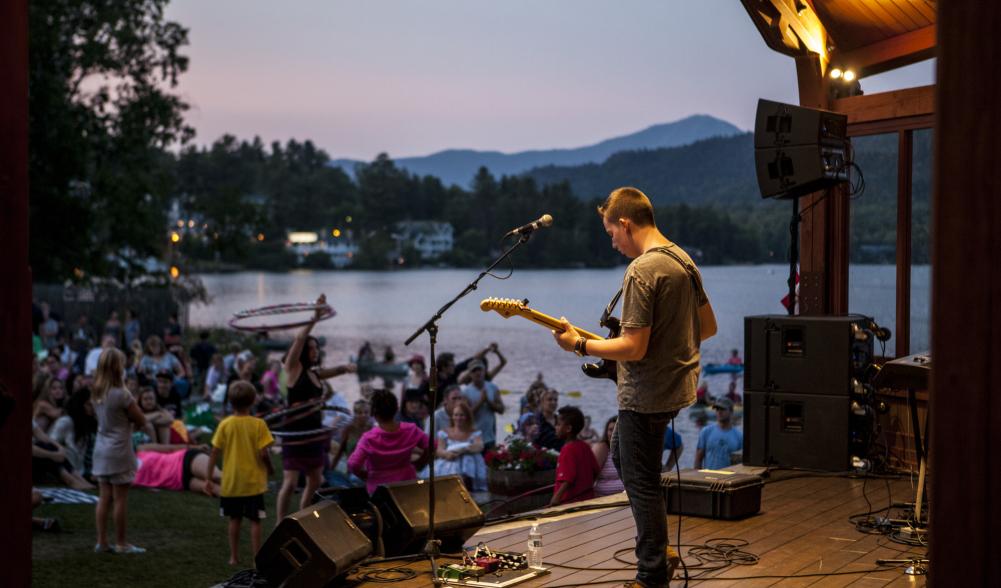 A man playing guitar on a stage next to a lake with people dancing in the background.