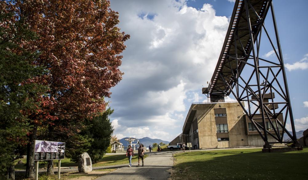 A couple walks on a path near a tree with red leaves and the towering Olympic ski jump.
