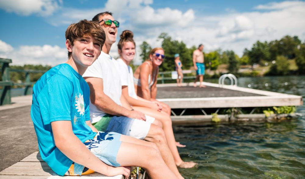 Two adults and a group of teenagers sit in swimming clothing on the edge of a dock in a lake.