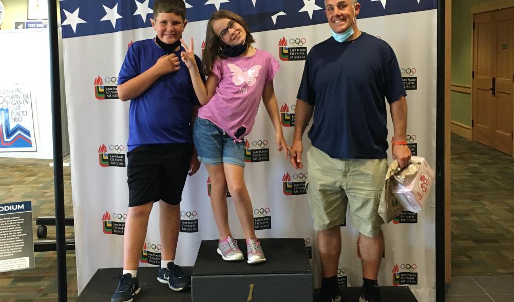 Two children and an adult male stand on a pretend Olympic medal stand in front of a stars and stripes backdrop.