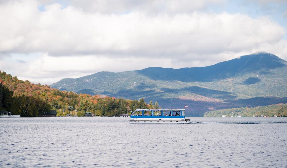 A tour boat cruises on a sparkling blue lake with fall foliage-covered mountains in the background.