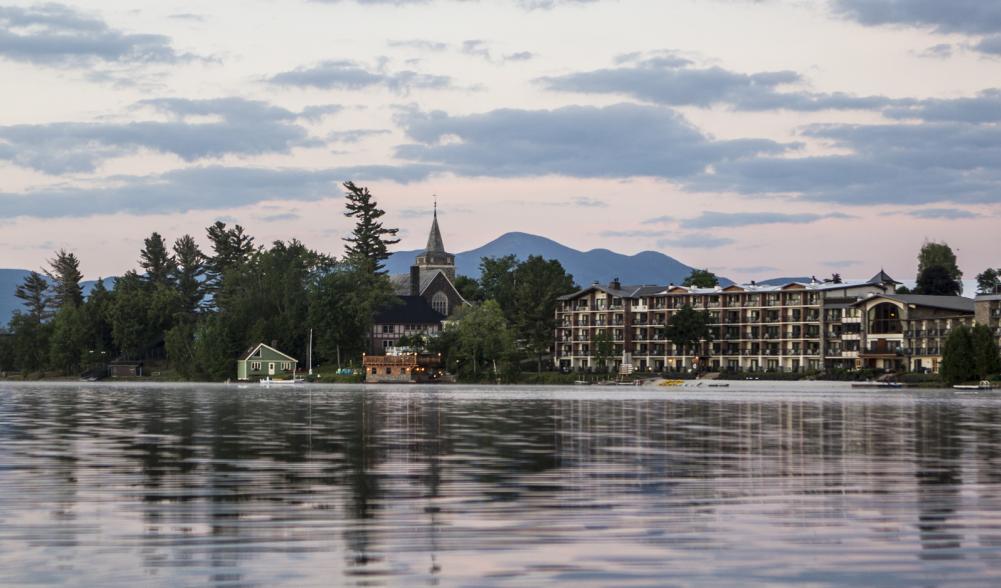 A wide hotel, church, and other buildings amid trees and a backdrop of mountains with a lake in the foreground.