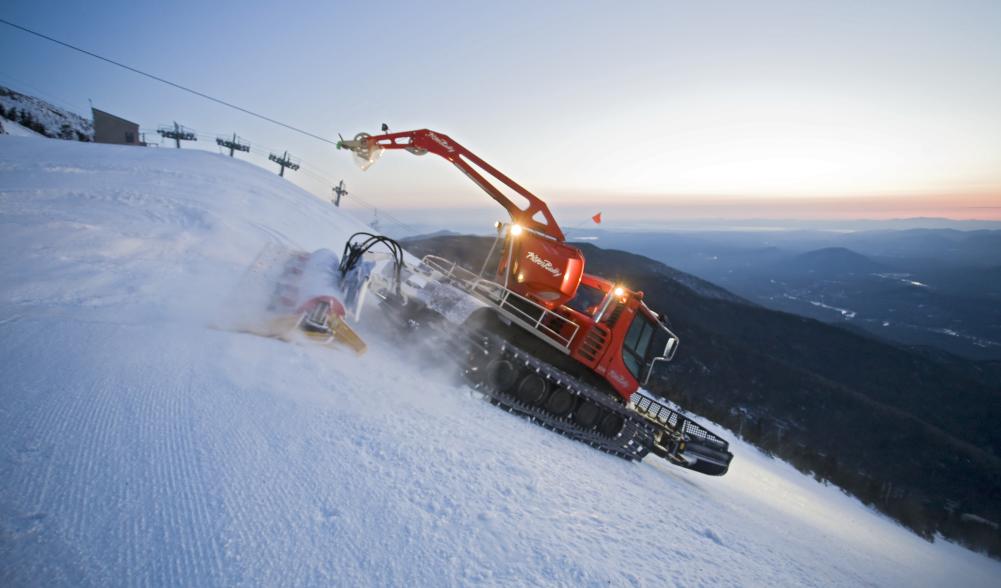 A large snow-grooming machine works its way down a snowy mountain.