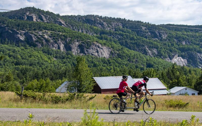 Tandem cyclists on road with barn and cliffs in background