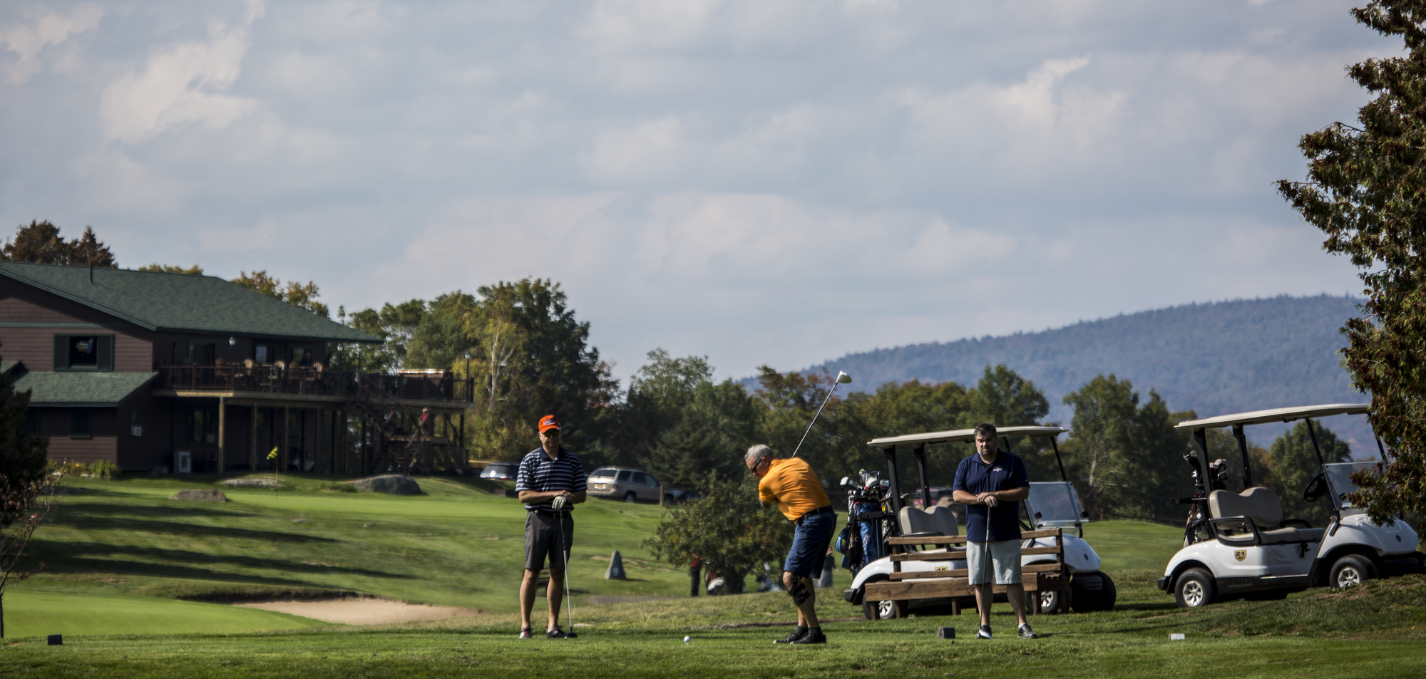 Three men golf with golf carts and clubhouse in the background.