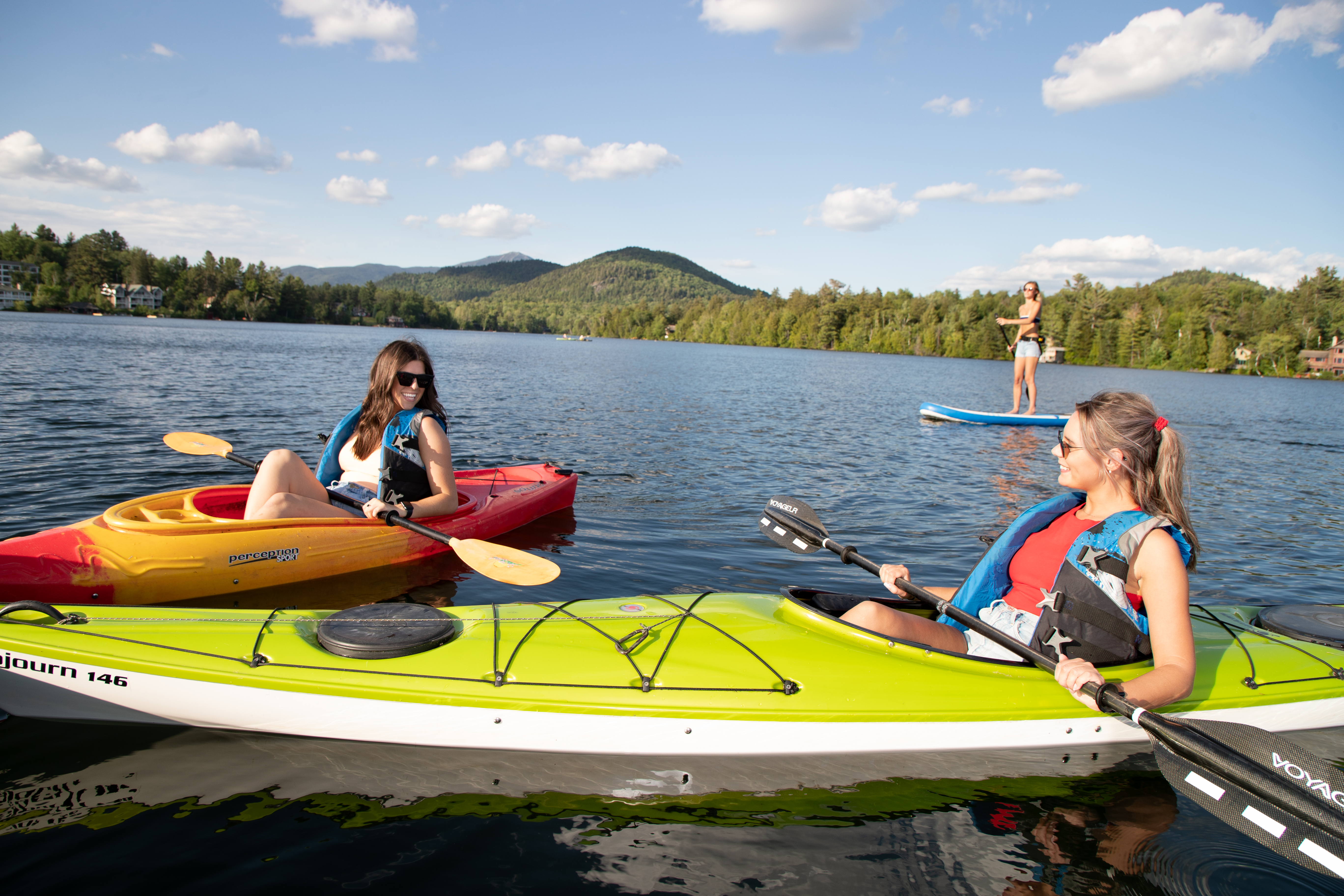 Two women kayak and another paddles a SUP on Mirror Lake&#44; all of which can be found at Lake Placid boat rentals.