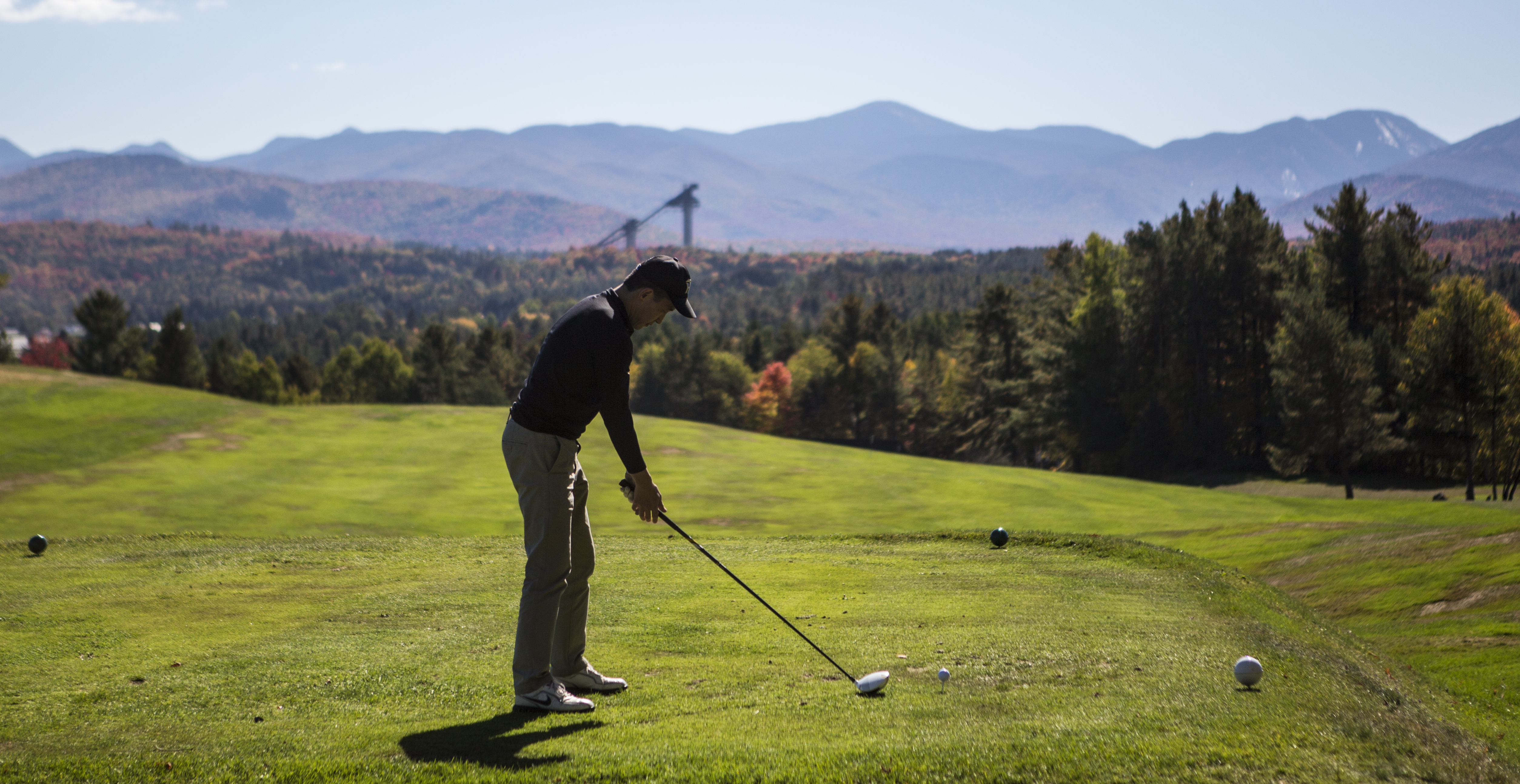 A golfer tees off with the Adirondack High Peaks and Olympic ski jumps in the background.
