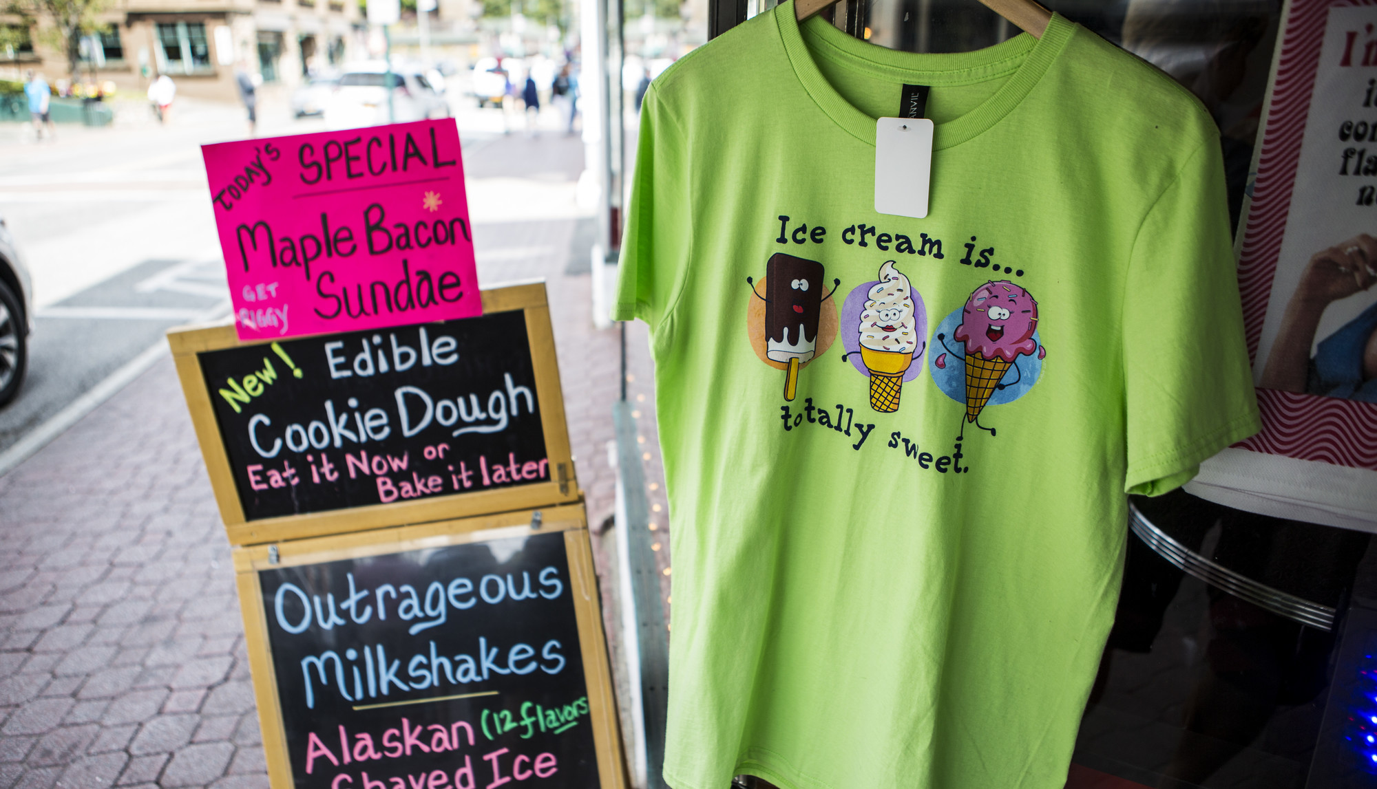 A sign advertising maple bacon ice cream sundaes sits next to a tshirt outside a shop.