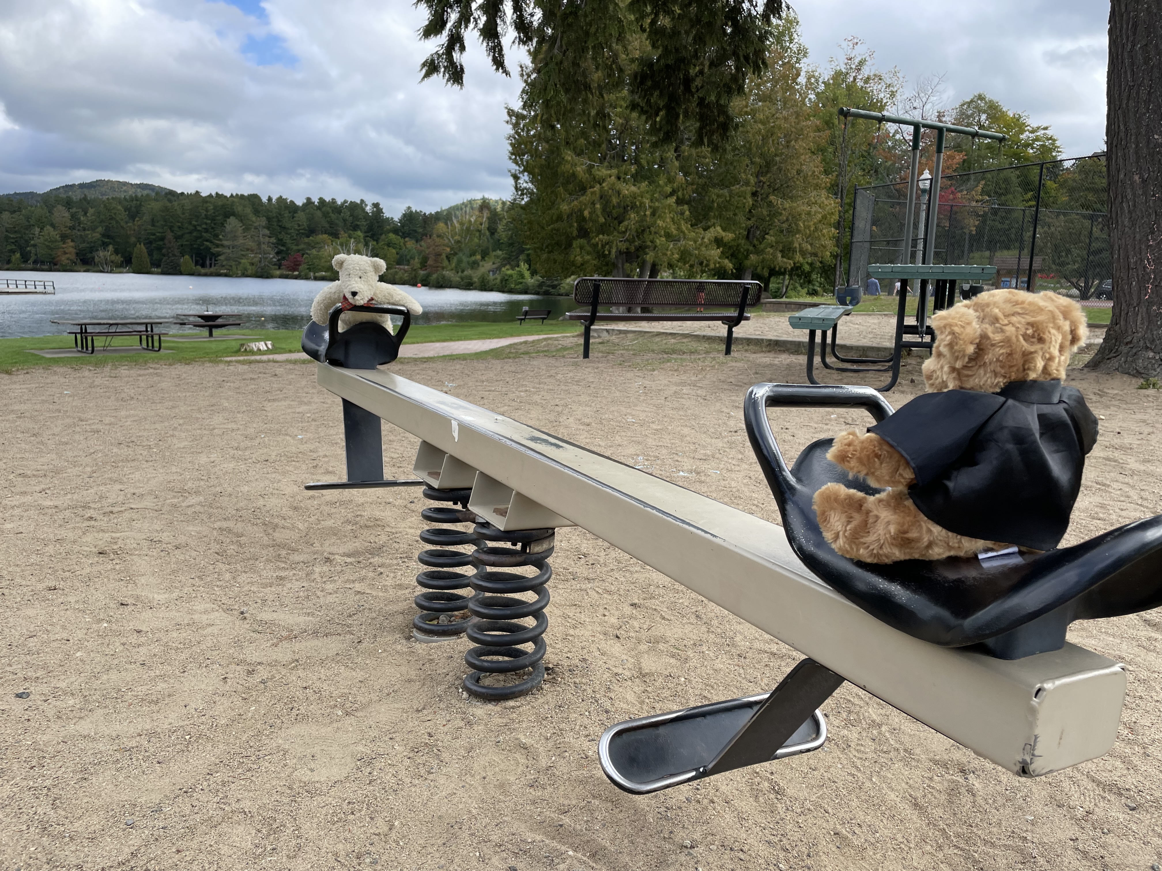 Two teddy bears sit on a teeter totter on a playground with a lake in the background.