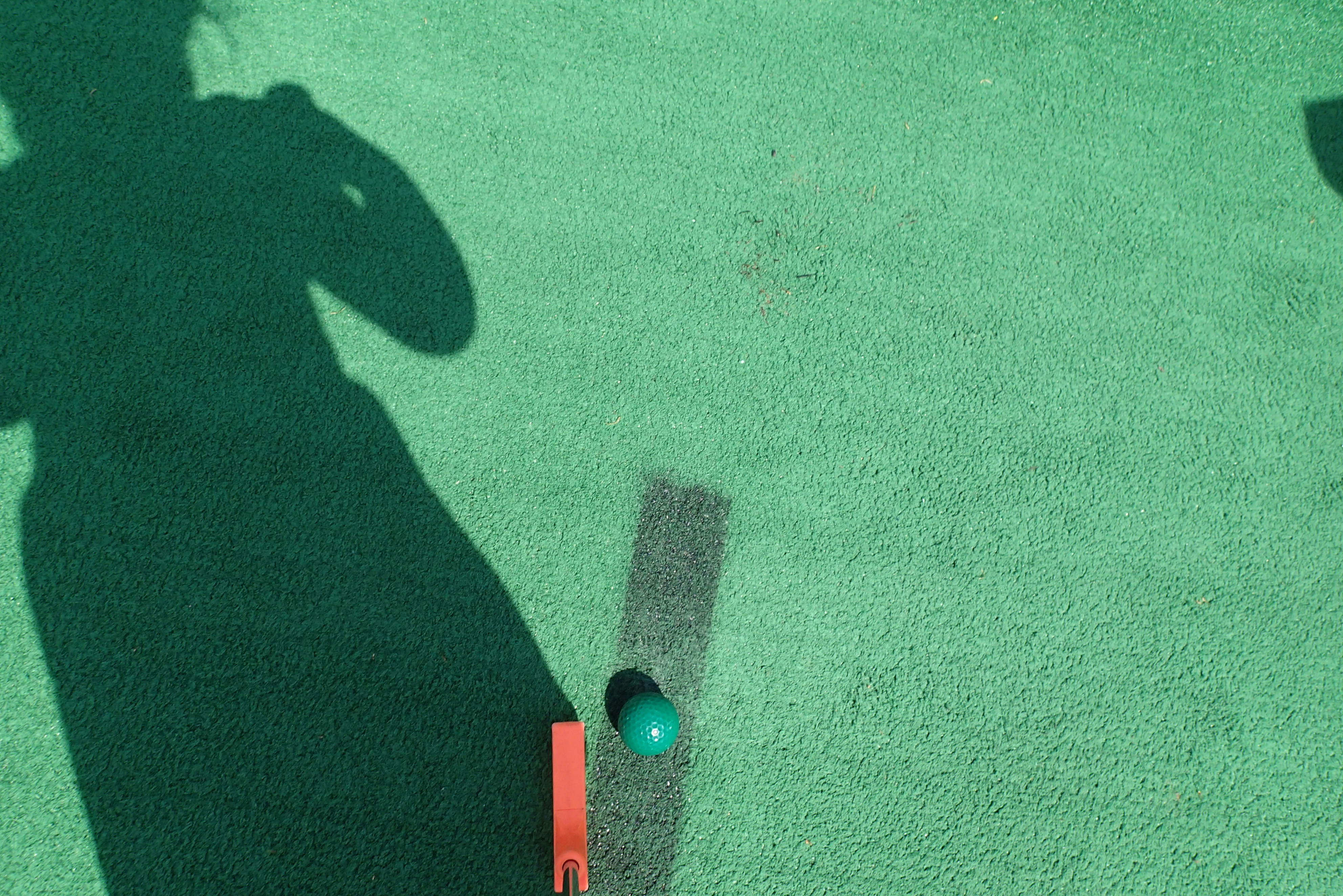 An image of artificial turf on a mini golf course&#44; with a putter&#44; golf ball&#44; and the shadow of a little girl.