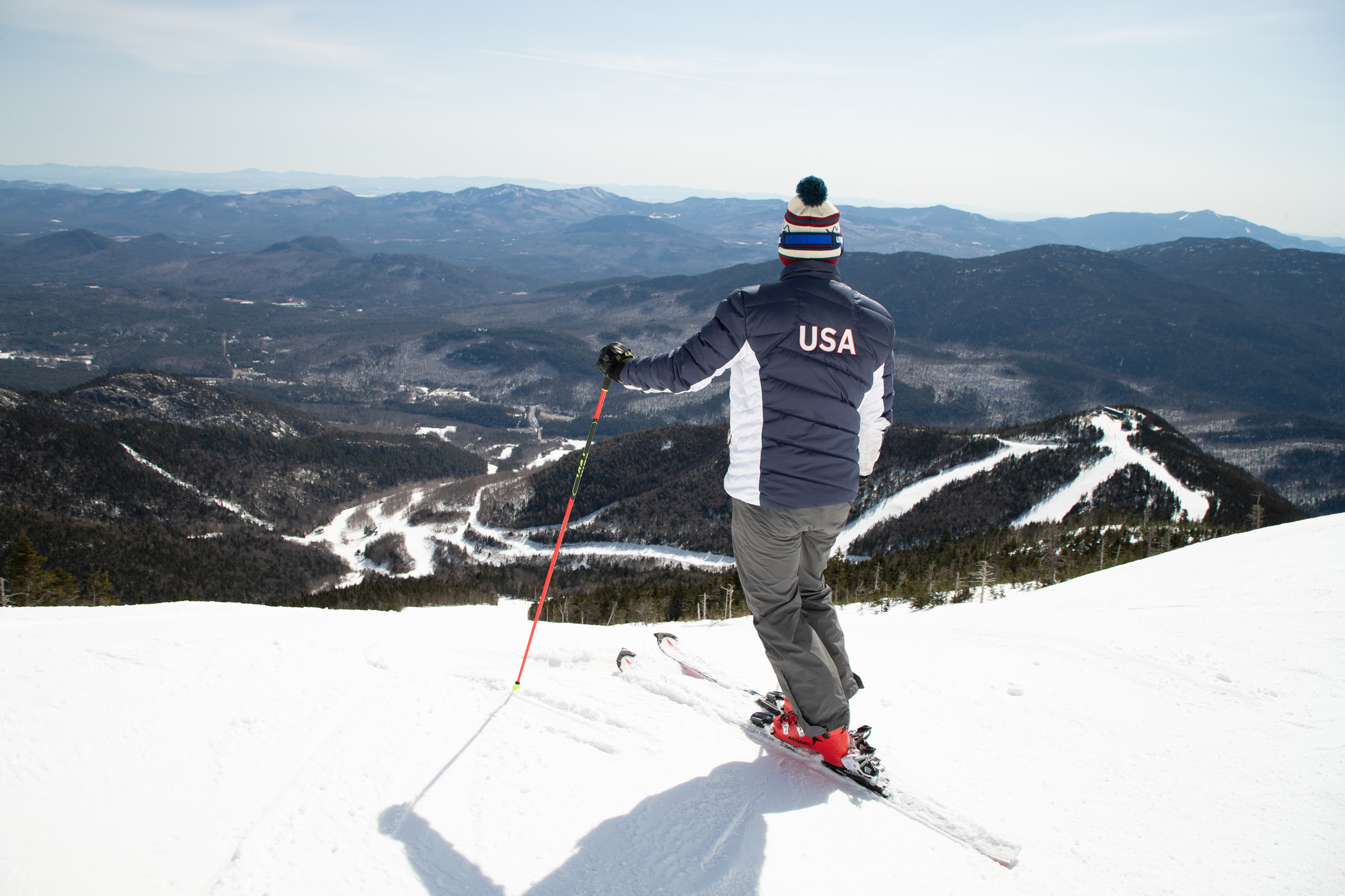 A skier in a red&#44; white&#44; and blue USA ski jacket poses on the edge of a run on a snowy mountain peak.
