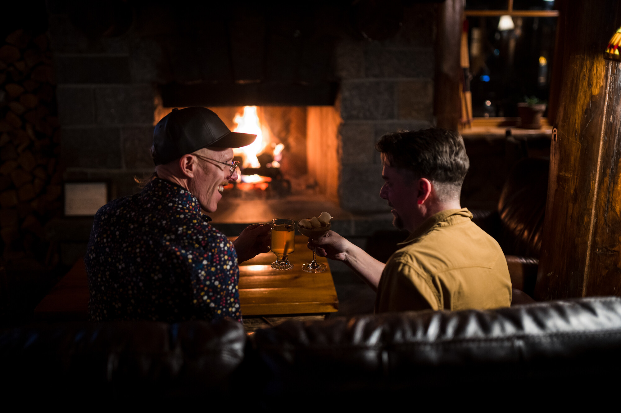 Two men cozy up by the fire with drinks in a dimly lit restaurant.