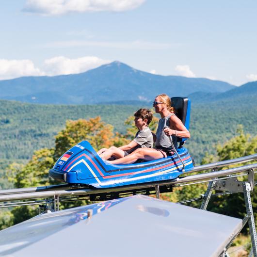 A woman and son ride in a roller coaster car on a track overlooking green mountains.
