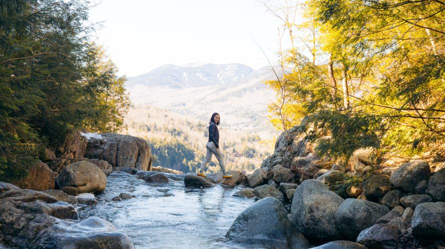 A woman walks across rocks on a stream overlooking the mountains.