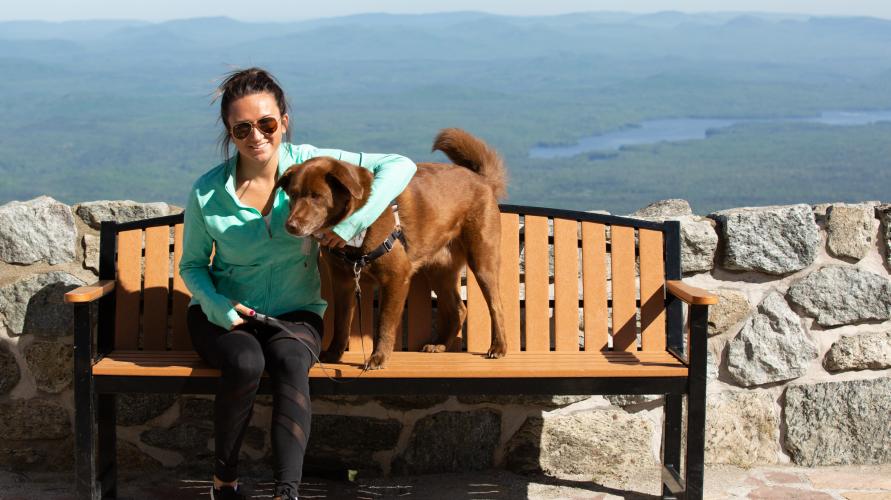 A woman sits on a bench with her dog overlooking the surrounding Adirondacks