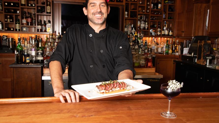 a chef holds a plate of tuna and rice at a bar with a cocktail on the side. 