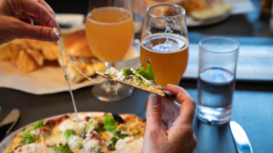A close up of a hand holding a cheesy piece of pizza with glasses of craft beer on the table next to the pizza.