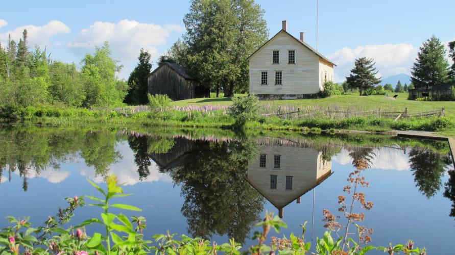 A rustic, historic farmhouse sits alongside a calm pond on a sunny day.