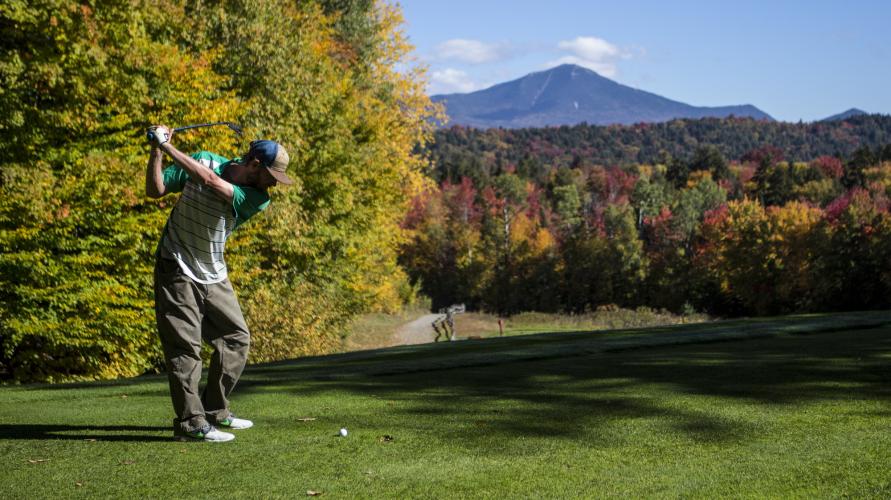 A man hits a golf ball in front of fall foliage