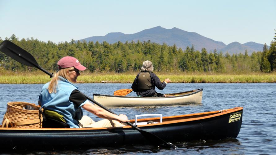 A man and woman in classic canoes paddle in front of High Peaks and marshland. 
