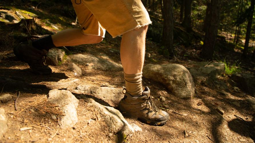 Close up of the lower half a person in shorts and hiking boots on a dirt and rock trail.