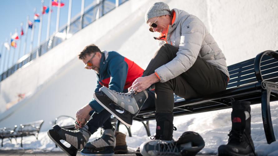 Two people strap on ice skates on a bench