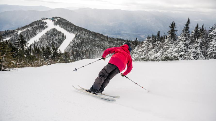 A skier carves down a snowy slope towards a sub-summit of a ski mountain.