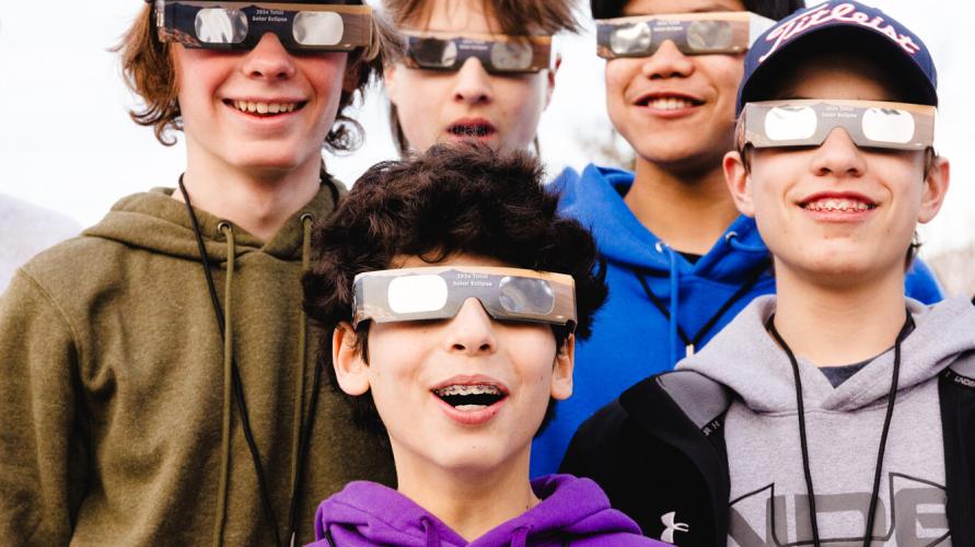 A group of five teenagers smile while wearing eclipse glasses.