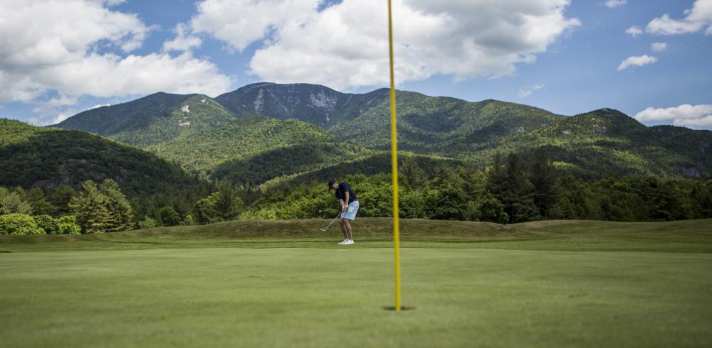 A man hits a golf ball into a hole with mountains in the background.