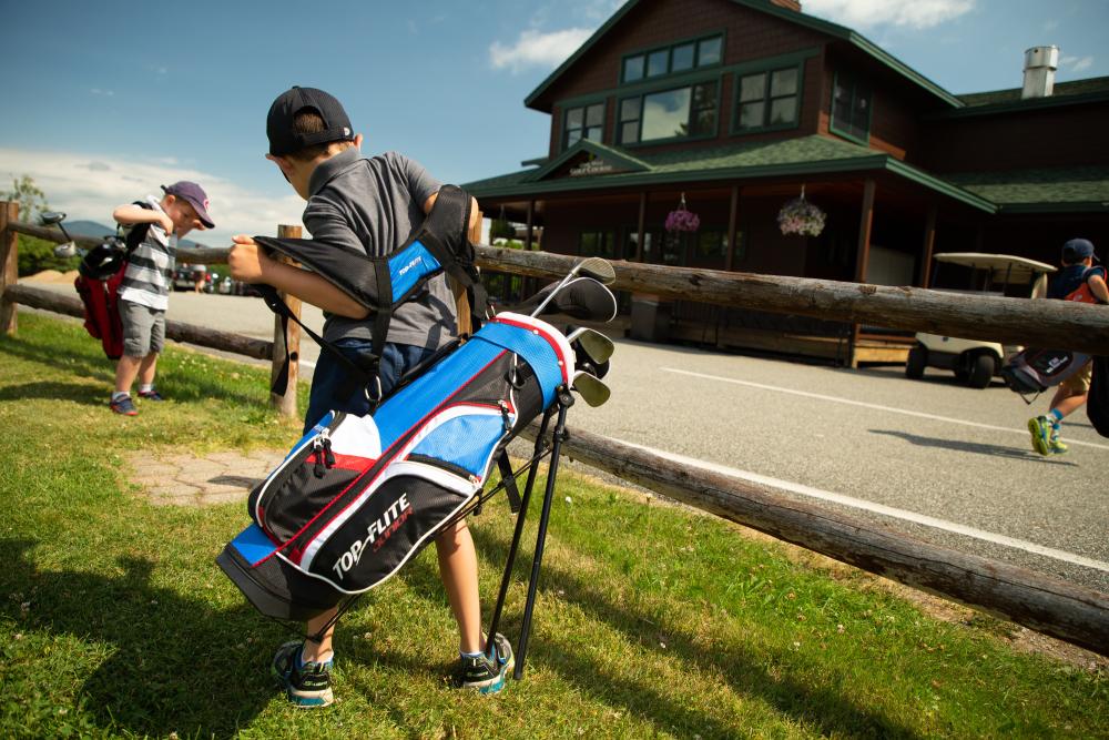 two boys shrug on golf club carriers in front of a golf course lodge.