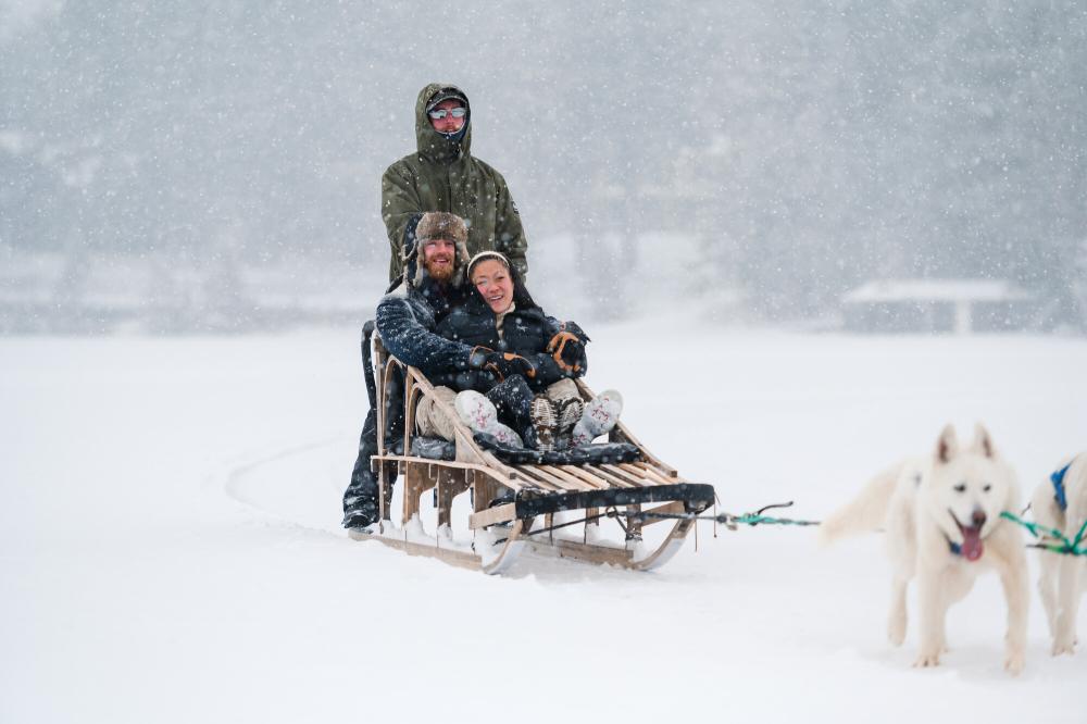 Couple smiles as they enjoy a ride in a dog. Heavy snow on Mirror Lake in Lake Placid.